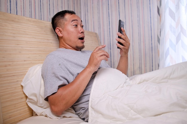 Happy excited Asian man sitting on bed at home using cell phone reading good news