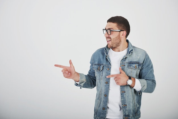 Happy excited bearded man in eyeglasses looking at camera with smile and pointing away at copy space with two fingers isolated over white background.