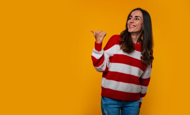 Happy and excited lovely smiling young woman in a sweater while she pointing away at advertising and isolated on yellow background