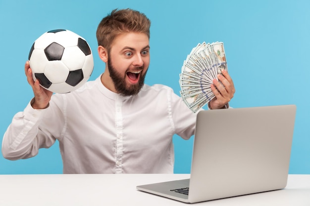 Happy excited man with beard holding fan of dollar cash and soccer ball looking at laptop display, betting and winning jackpot. Indoor studio shot isolated on blue background