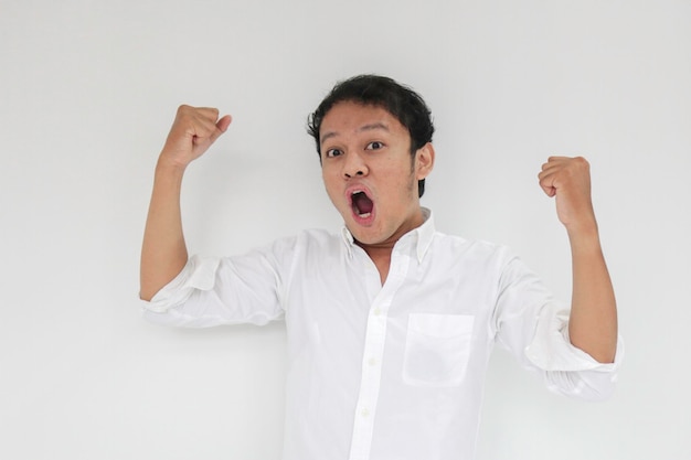 Happy excited and smiling young Asian man raising his arm up to celebrate success or achievement Indonesian man wearing white shirt
