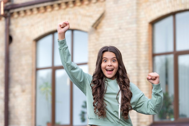 カジュアルなスタイルのぼやけた屋外で勝者のジェスチャーを作る幸せな興奮した 10 代の少女