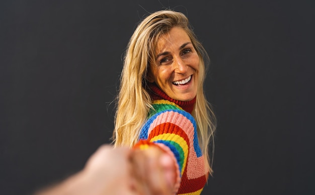 Happy excited woman pulling guy's hand wearing colorful rainbow sweater standing over black background lgbt celebrating concept image