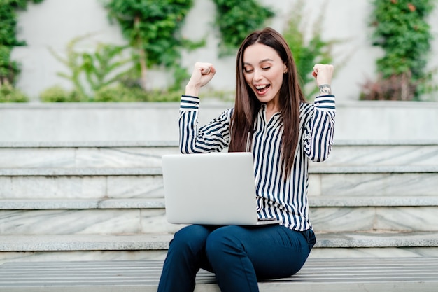 Happy and excited woman sitting with laptop outdoors