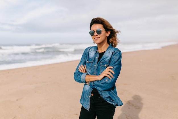 Happy excited woman with flying hair wearing denim shirt and sunglasses is posing on the beach with ocean background in sunlight Travel woman walking on the ocean shore in warm spring day