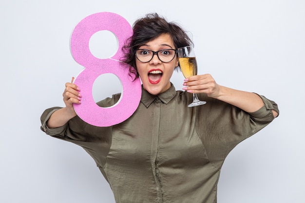Счастливая и взволнованная женщина с короткими волосами, держащая номер восемь из картона и бокал шампанского, весело улыбается, празднуя международный женский день 8 марта