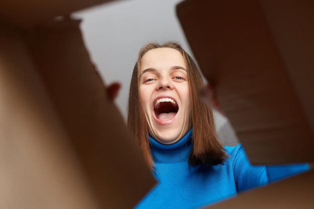 Счастливая и взволнованная молодая взрослая женщина, заглядывающая внутрь картонной коробки Выглядящая веселой и радостной, кричащей от счастья, хорошая служба доставки Вид снизу картонной коробки