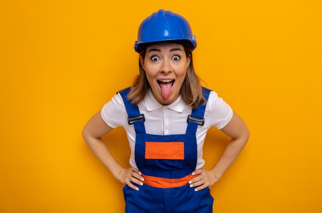 Happy and excited young builder woman in construction uniform and safety helmet sticking out tongue standing over orange wall