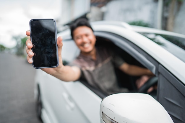 Счастливый возбужденный молодой мужчина-водитель показывает экран своего мобильного телефона, сидя в машине