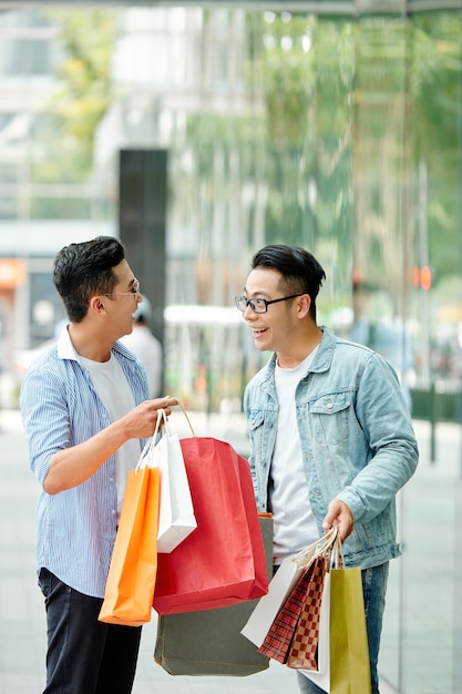 Happy excited young Vietnamese men discussing purchases they made on sale in shopping mall