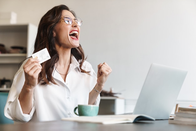 a happy excited young woman posing indoors using laptop computer holding credit card.