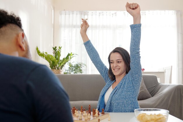 Happy excited young woman raising arms and celebrating victory in game of chess with her boyfriend