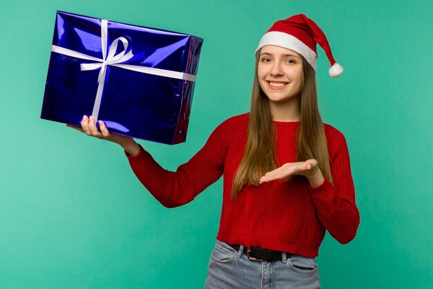 파란색 배경 위에 선물 상자가 있는 산타클로스 모자를 쓴 행복한 흥분된 젊은 여성 - 이미지