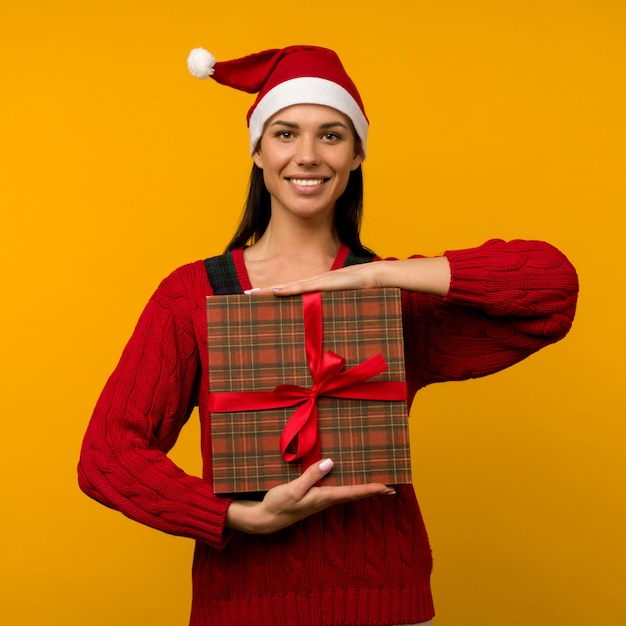 Felice eccitato giovane donna in cappello di babbo natale con confezione regalo su sfondo giallo - immagine