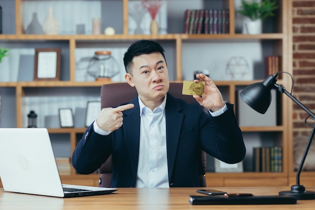 Happy and exciting asian banking businessman looking at camera
and smiling working in classic office holding thumbs up and
offering gold bank credit card