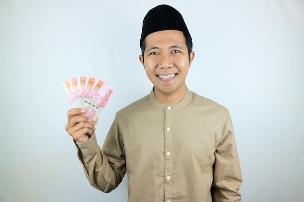 Foto espressione felice di un uomo musulmano asiatico che tiene in mano banconote di rupiah isolate su sfondo bianco