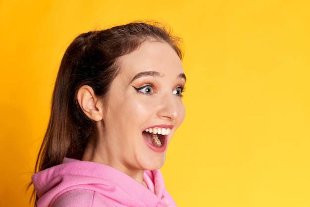 Happy extremely excited Portrait of young beautiful girl posing in pink hoodie over yellow studio background Concept of youth emotions