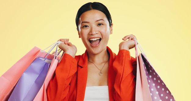 Happy face and woman in studio with shopping bag and boutique sale on yellow background Portrait excited and asian girl shopper customer with items for discount fun and retail while isolated
