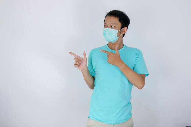 Happy face of young Asian man wear medical mask and blue with hand point on empty space