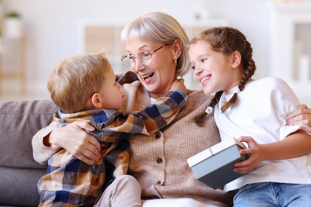 Photo happy family aged woman grandmother laughing and embracing grandkids boy and girl with present while resting on sofa during holiday celebration at home
