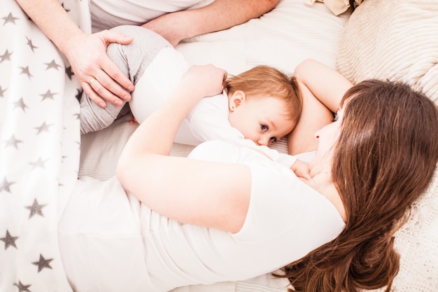 Happy family on the bed. Mother and farther sleep with their little girl. Mom is breastfeeding her toddler daughter