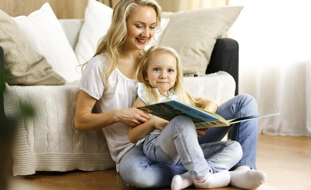Фото Счастливая семья. блондинка молодая мать читает книгу своей милой дочери, сидя на деревянном полу. концепция материнства.