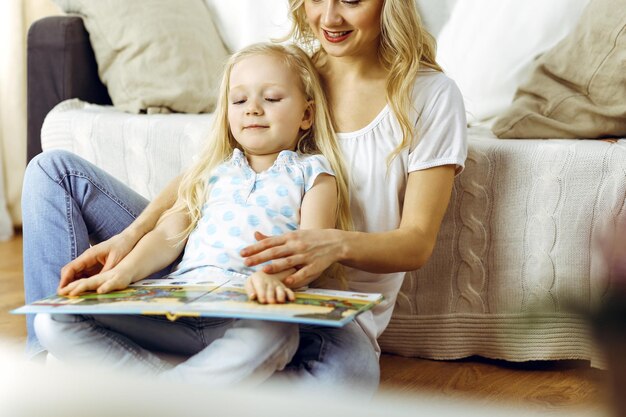 Happy family. Blonde young mother reading a book to her cute daughter while sitting at wooden floor. Motherhood concept