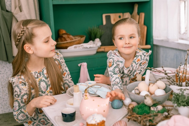 幸せな家族の率直な小さな子供姉妹の女の子は一緒に楽しい準備ができて、ランチやディナーにイースターケーキベーカリーとキャンディーが付いたキッチンで飾られたテーブルで春のイースター休暇を待っています