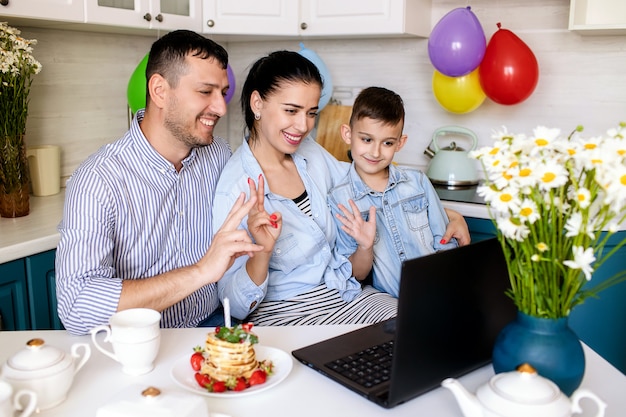 Happy family celebrating a birthday at home in the kitchen and chatting online on a laptop