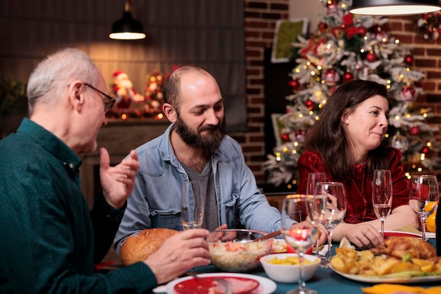 クリスマスを祝う幸せな家族、お祝いの夕食の席で話し、伝統的な冬の休日の食べ物を食べ、おしゃべりをする人々。ホームパーティーで両親とクリスマスイブのお祝い