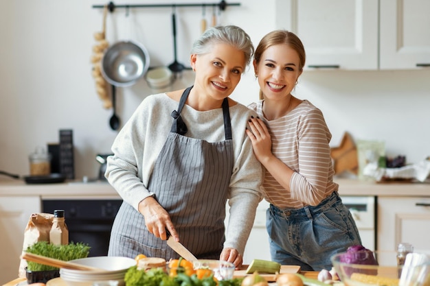 Happy family cheerful young woman embracing mature mother while preparing healthy dish with fresh vegetables in home kitchen