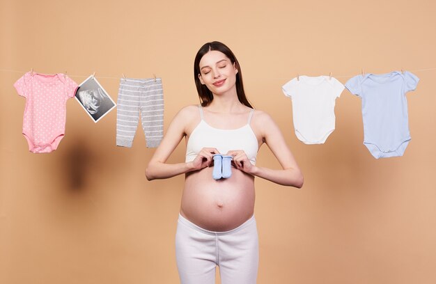 Happy family, childcare concept, pregnancy, solo parenting. Young happy pregnant woman, holding infant blue socks, waiting for her son, on a rope in the background, dries children's clothes.