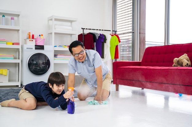 幸せな家族が部屋を掃除します。アジア人の父と息子が家の掃除をします。若い男と子供の女の子が居間でほこりを払い、タオルとスプレーで床を洗っています