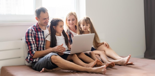 행복한 가족 개념입니다. 아름다운 어머니와 잘생긴 아버지는 딸들과 함께 집에서 함께 시간을 보내고 노트북을 들고 침대에 누워 있습니다.