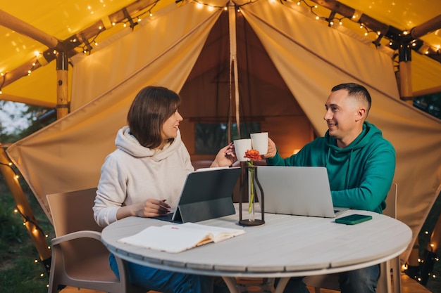Happy family couple freelancers drink coffee while working\
laptop on cozy glamping tent in summer evening luxury camping tent\
for outdoor holiday and vacation lifestyle concept