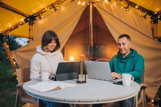 Felice coppia di famiglia freelance che lavorano laptop su un'accogliente tenda glamping in serata estiva tenda da campeggio di lusso per vacanze e vacanze all'aperto concetto di stile di vita