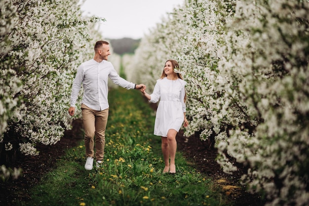 Happy family couple in spring blooming apple orchard. Young couple in love enjoy each other while walking in the garden. The man holds the woman's hand. Family relationships