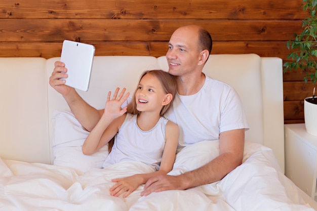 Happy family, dad and daughter talking on the tablet at home in bed. Distance learning.