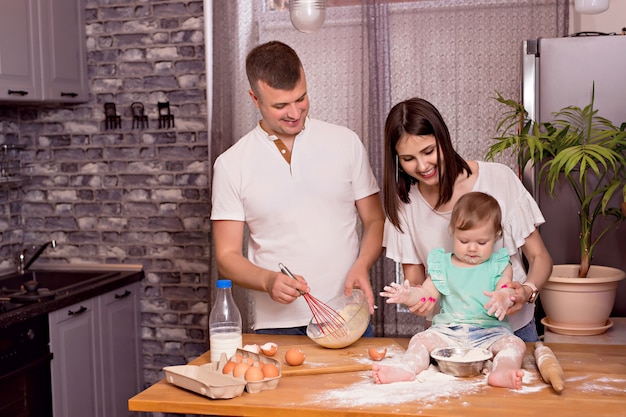 Famiglia felice, papà, mamma e figlia giocano e cucinano in cucina, impastano e cuociono i biscotti