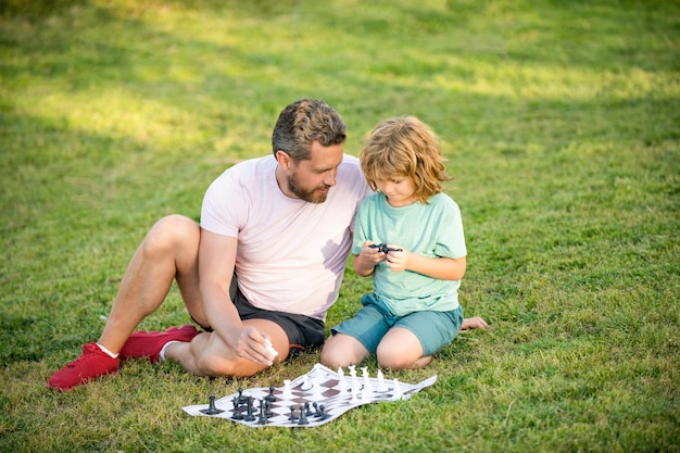 Happy family of daddy and son kid playing chess on green grass\
in park outdoor, decision