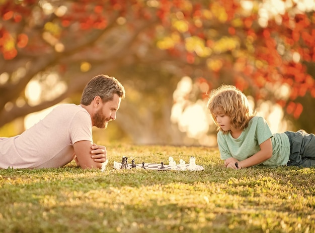 Happy family of daddy and son kid playing chess on green grass in park outdoor genius