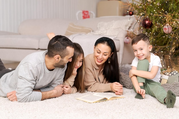 Happy family in decorated house at christmas lying on floor reading book against background of couch