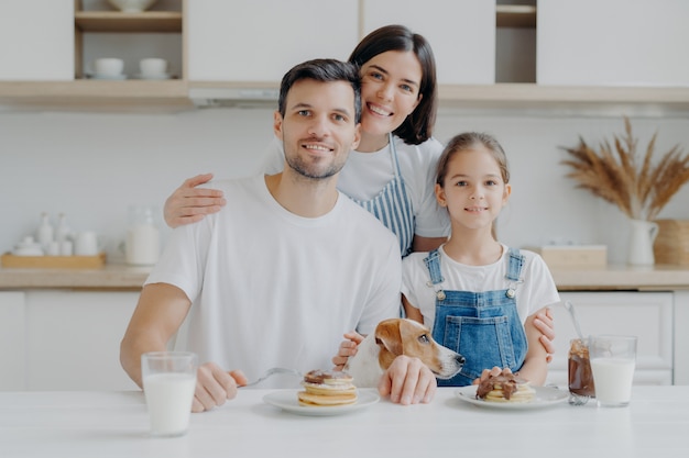 Happy family and dog pose in cozy kitchen, eat fresh homemade pancakes with chocolate and milk, look positively at camera. Mother in apron embraces husband and daughter, likes cooking for them