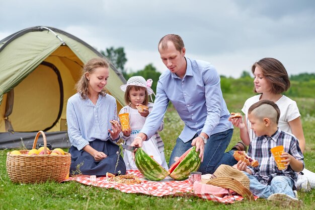 Happy family eating watermelon at picnic in meadow near the tent Family Enjoying Camping Holiday In Countryside