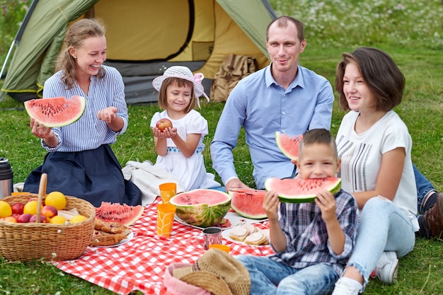 テントの近くの牧草地でピクニックでスイカを食べる幸せな家族。田舎でキャンプ休暇を楽しんでいる家族。