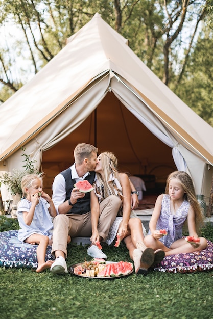Счастливая семья ест арбуз на пикнике на лугу возле палатки вигвам типи