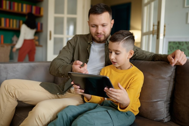 Happy family enjoy fun time together on weekend at home. Father and son embracing while using mobile tablet while mother reading book