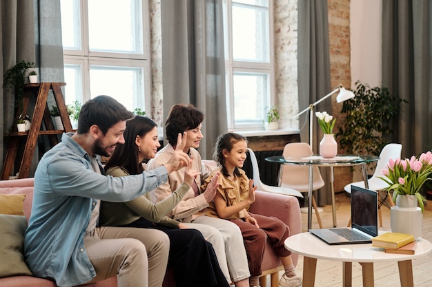 Happy family of four sitting in row on large soft comfortable couch in living room and communicating with their friends on laptop display