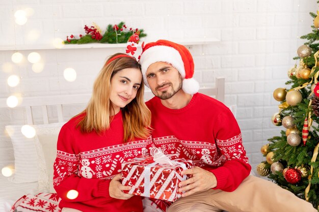 Happy family guy and girl in red sweaters under the Christmas tree on the bed at home celebrate New Year or Christmas rejoicing and giving each other gifts