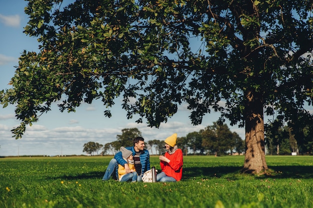 Счастливая семья устраивает осенний пикник, сидит на зеленой траве, пьет горячий чай, общается друг с другом